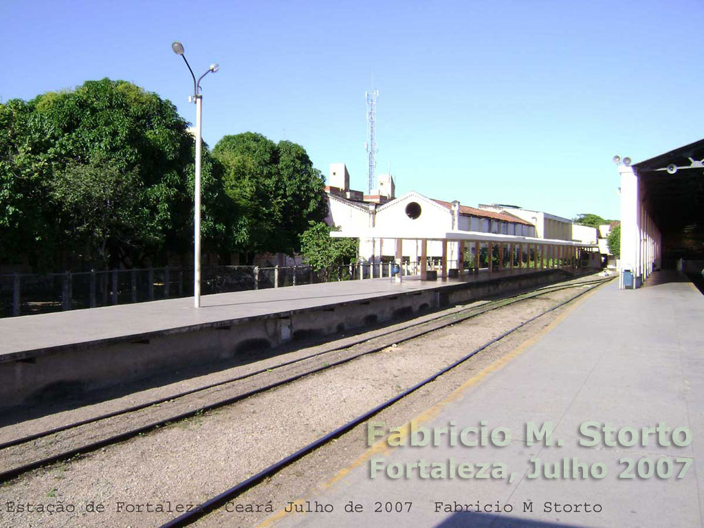 Trilhos e plataforma do trem urbano, vendo-se à direita a gare de passageiros e à esquerda prédios das oficinas ferroviárias